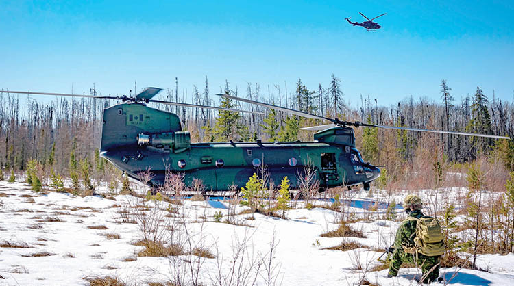 Side view of chinook helicopter on snowy ground, forest in background, griffin helicopter in sky on right side