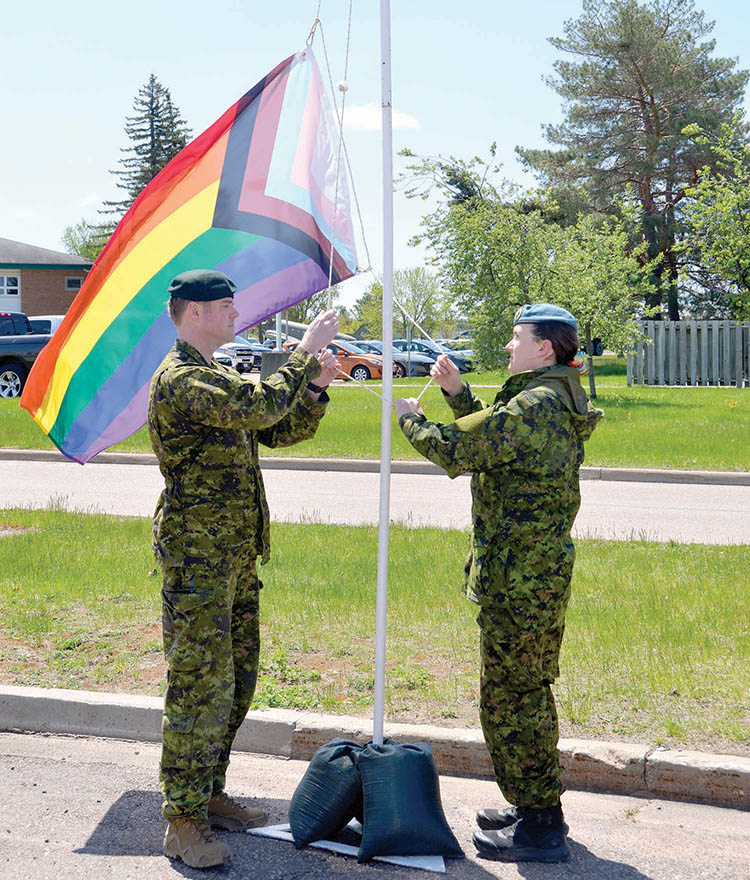 A male and female soldier stand on either side of a flag pole raise pride flag