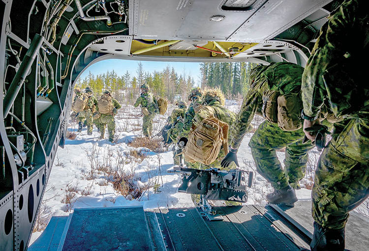 Photo taken from inside helicopter, several soldiers disembarking through back into snow covered field, each soldier carries a rucksack